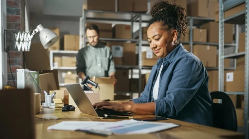 A smiling woman works on a laptop while a coworker packs boxes on shelves.