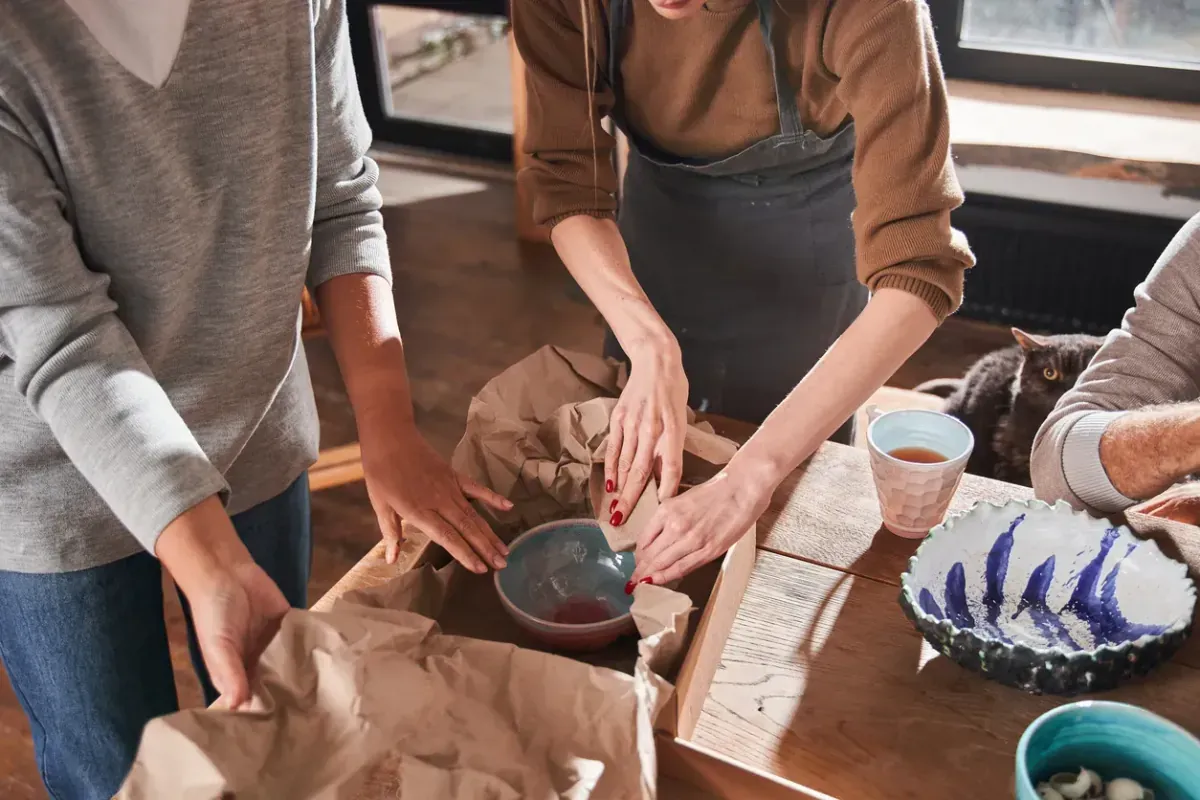 Cropped view of the two women putting a new handmade plate on the paper and in the cupboard box while packing it.