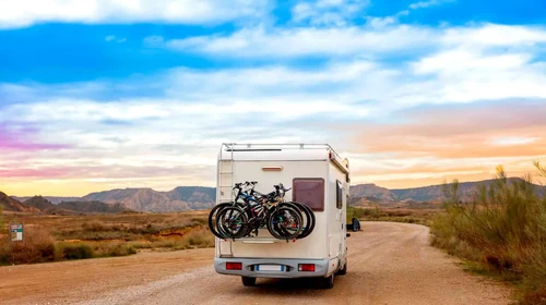 A camper van carrying bikes driving along a desert dirt road at sunset.