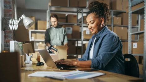 A smiling woman works on a laptop while a coworker packs boxes on shelves.